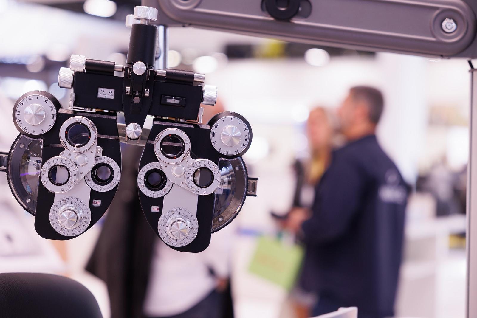 A phoropter in the foreground of an instruments stand, visitors out of focus behind it.